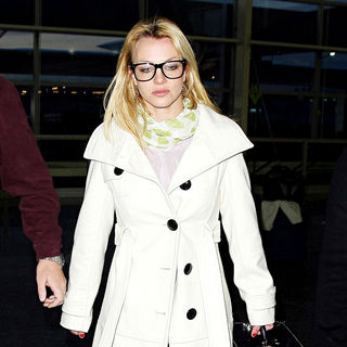 Britney Spears wearing large black rimmed glasses and a white coat catches a flight